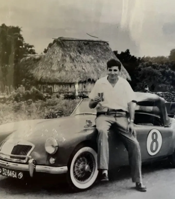 Fotografía antigua: un joven apoyado en un MG descapotable junto a una palapa
